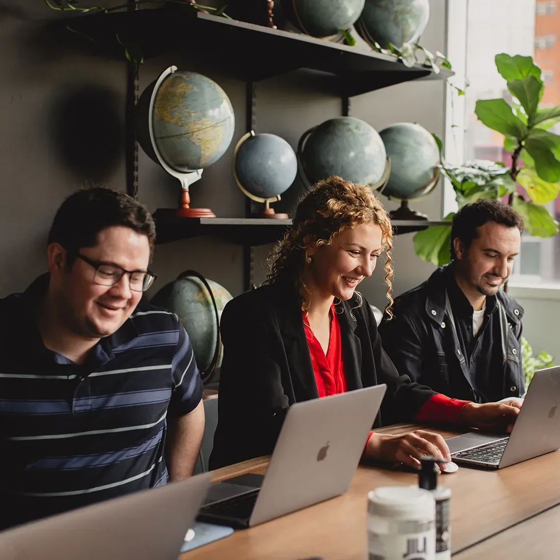 Three BFJ Digital team members smiling as they work on laptops at a shared desk in the office