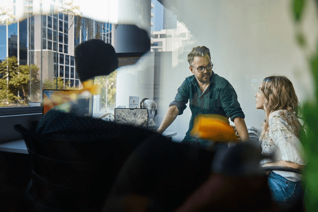Two members of the BFJ Digital marketing team talking at a desk in the Brisbane office, seen through the glass with city buildings outside