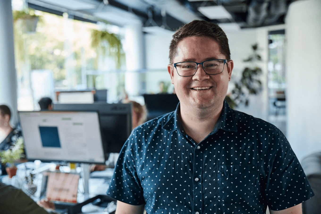 A smiling BFJ Digital team member in the open-plan Brisbane office, with colleagues working at their desks behind