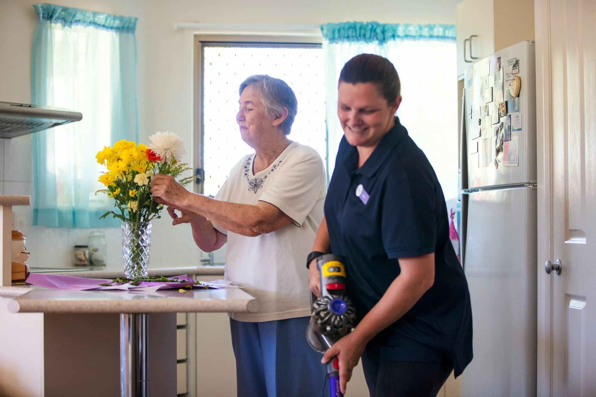 A older woman in the kitchen with a lady helping her clean it