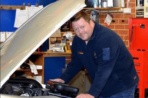 A mechanic standing in front of a car with the hood propped up